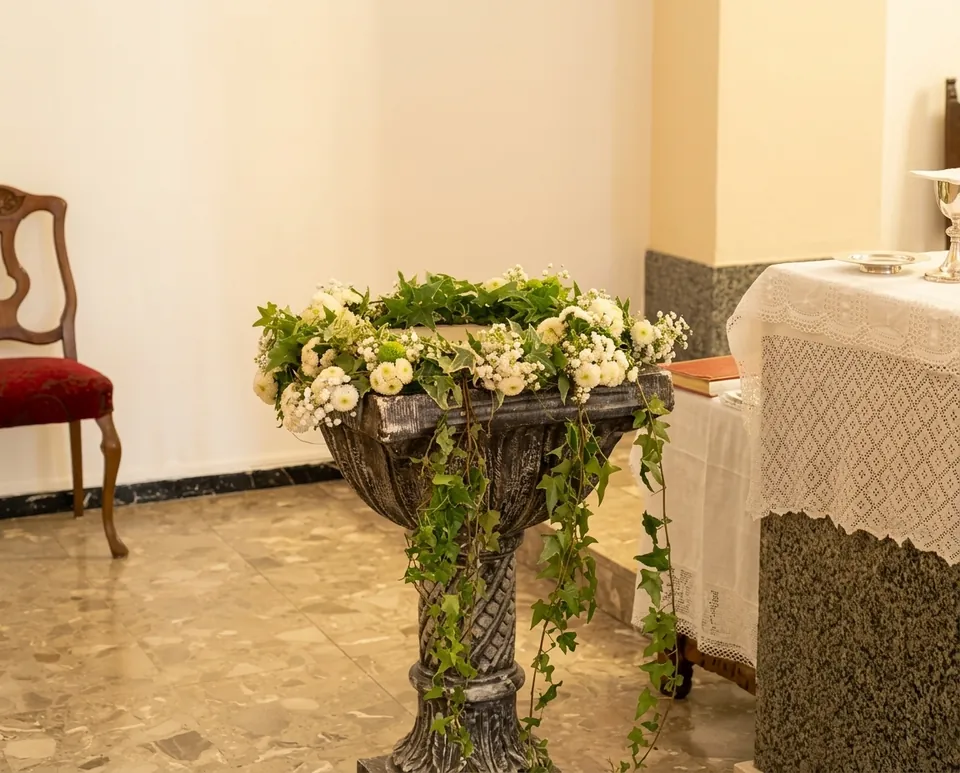 Copa de piedra decorada con flores blancas, crisantemos verdes e hiedra colgante junto al altar de una iglesia, preparada para un bautizo