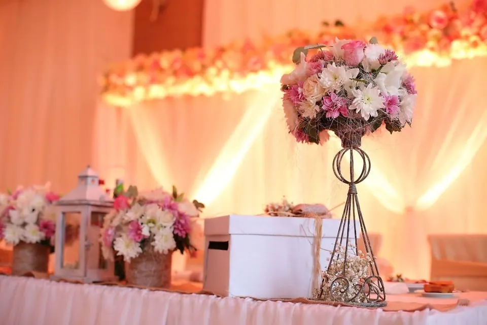 Centro alto de flores rosas y blancas sobre la mesa de una celebración de comunión, con farolillos y guirnalda floral al fondo